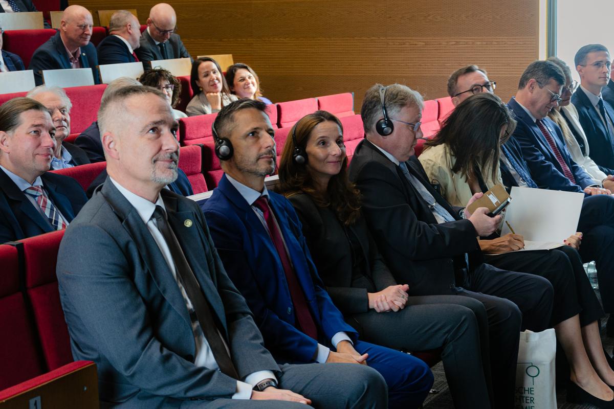 Przedstawia fragment publiczności podczas Uroczystej Inauguracji objęcia stanowiska przez Dyrektora Piotra Girdwoynia. Na pierwszym planie znajduje się Zastępca Dyrektora kierujący Ośrodkiem Aplikacji Prokuratorskiej prokurator Andrzej Piaseczny. Obok niego siedzą w słuchawkach zagraniczni goście. Jedna z kobiet w pierwszym rzędzie coś notuje. 