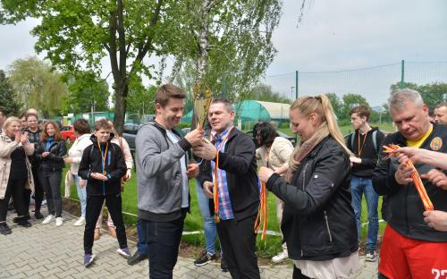 Zdjęcie nr 19: mężczyzna z medalami w ręce, obok niego kobieta również z medalami, obok niej mężczyzna wręczający puchar zawodnikowi, wydarzenie obserwuje grupa osób, w tle stadion sportowy