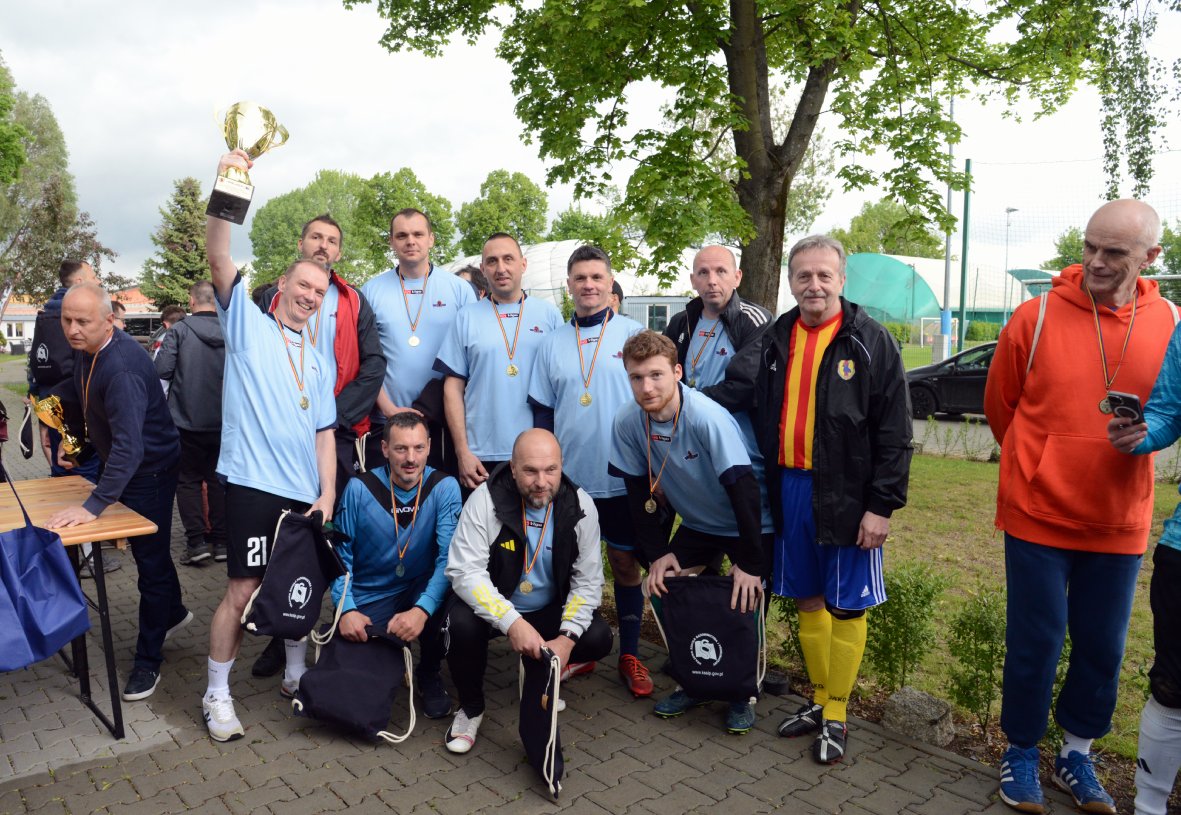 Na zdjęciu grupa kilkunastu mężczyzn pozuje wspólnie na tle zielonych drzew i sportowego boiska. Większość z nich nosi jasnoniebieskie koszulki sportowe, a na szyjach mają zawieszone złote medale. Jeden z zawodników unosi w górę duży, złoty puchar. Kilku uczestników klęczy z przodu, trzymając granatowe worki sportowe. Po prawej stronie stoi mężczyzna w kurtce w czerwono-żółte pasy, niebieskich spodenkach i żółtych getrach, a obok niego osoba w czerwonej bluzie z telefonem w ręku. W tle widać zielone drzewa, namioty sportowe i samochód zaparkowany przy ulicy. Cała scena rozgrywa się na brukowanym placu pod rozłożystym drzewem