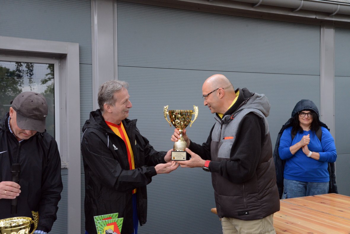 Na zdjęciu widać moment wręczenia pucharu podczas zawodów sportowych. Dwóch mężczyzn stoi naprzeciw siebie – jeden przekazuje drugiemu złoty puchar z uchwytami po bokach. Obaj są ubrani w sportowe kurtki, a w tle znajduje się szara ściana budynku z dużym oknem. Po lewej stronie stoi mężczyzna z mikrofonem, a po prawej kobieta w niebieskiej bluzie, która wydaje się być zadowolona i klaszcze w dłonie. Na drewnianym stole widać jeszcze jeden puchar oraz kolorowe dyplomy lub nagrody. Cała scena ma charakter oficjalny i radosny, związany z nagradzaniem osiągnięć.