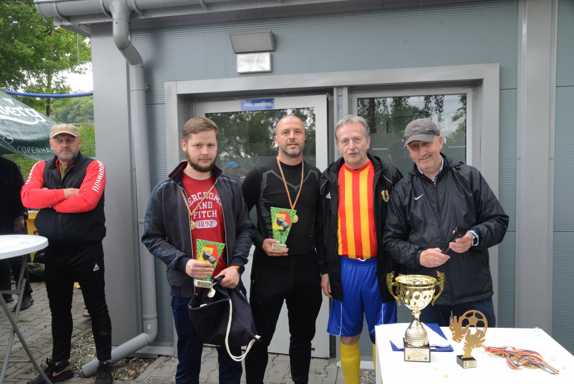 Na zdjęciu widać czterech mężczyzn stojących w rzędzie przed wejściem do budynku. Trzech z nich trzyma w rękach zielone statuetki z motywem sportowym, a jeden z nich ma na szyi złoty medal. Jeden z mężczyzn ubrany jest w sportowy strój: żółto-czerwoną koszulkę w pionowe pasy, niebieskie spodenki i żółte getry. Pozostali mają na sobie sportowe kurtki lub bluzy oraz codzienne ubrania. Po prawej stronie, na białym stole, ustawiony jest duży złoty puchar z uchwytami, mniejsza statuetka w kształcie piłki oraz kilka kolorowych medali na wstążkach. W tle widoczna jest szara ściana budynku i przeszklone drzwi. Cała scena ma charakter uroczysty i sportowy, związany z wręczaniem nagród.