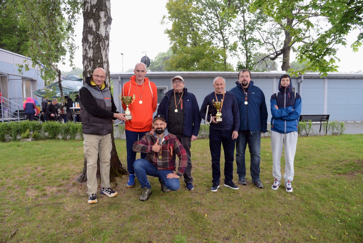 Na zdjęciu znajduje się grupa siedmiu mężczyzn pozujących na trawniku pod drzewem, na tle szarego budynku. Sześciu z nich stoi, a jeden klęczy z przodu i pokazuje uniesiony kciuk w geście radości. Kilku mężczyzn ma na szyjach złote medale na pomarańczowych wstążkach, a dwaj trzymają w rękach duże, złote puchary. Ubrani są w sportowe lub codzienne stroje – bluzy, kurtki, spodnie dresowe i sportowe buty. W tle widać zieleń drzew oraz kilka osób i stoły ustawione pod parasolami. Atmosfera jest radosna i uroczysta, związana z wręczeniem nagród po sportowej rywalizacji.