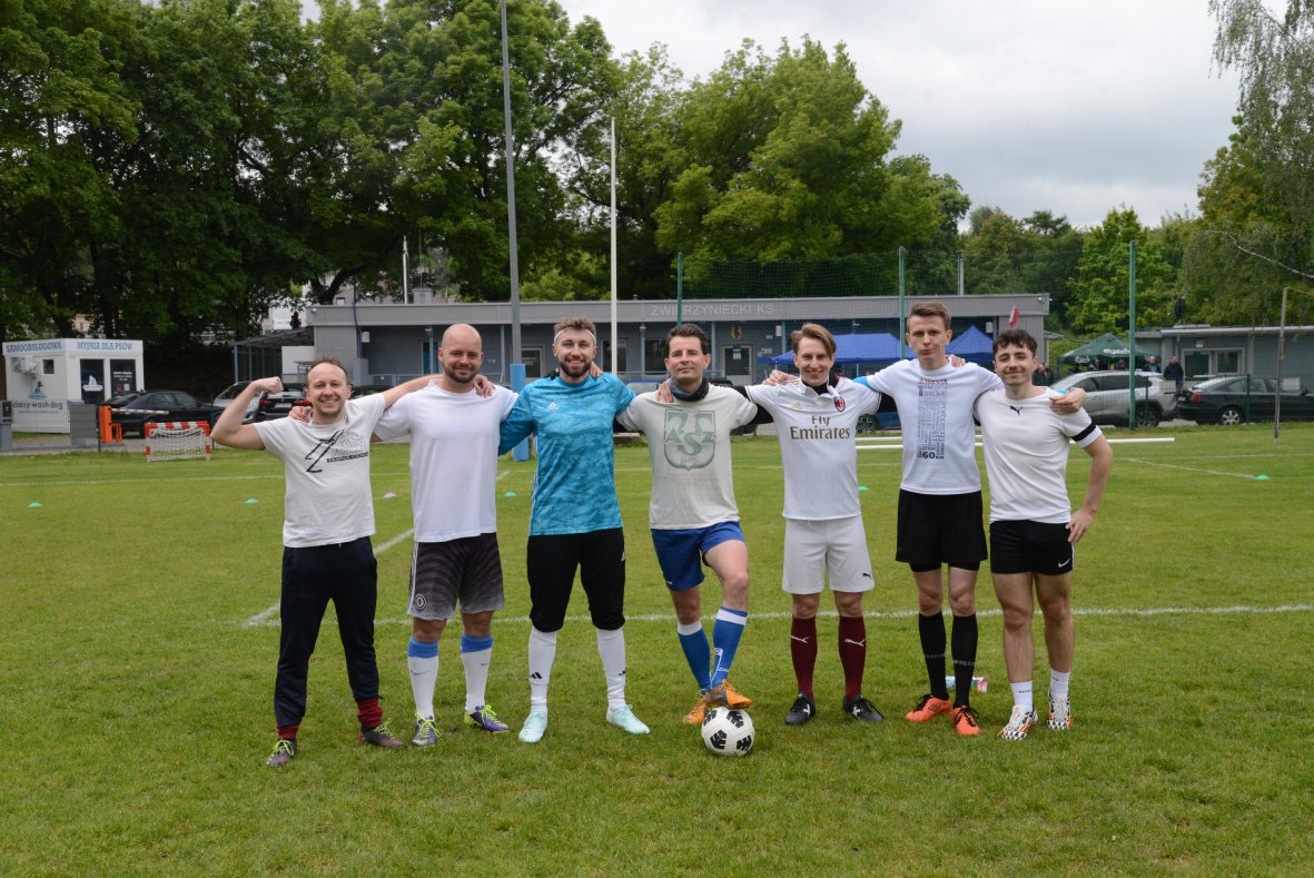 Na zdjęciu znajduje się siedmiu mężczyzn stojących ramię w ramię na murawie boiska piłkarskiego. Wszyscy są ubrani w sportowe stroje – dominują białe koszulki, choć jeden z mężczyzn ma na sobie niebieską bramkarską bluzę, a inny niebieskie spodenki i podniesioną nogę, opierając ją na piłce. Część osób ma na sobie kolorowe getry i buty piłkarskie. W tle widać zielone drzewa, budynki klubowe oraz zaparkowane samochody. Atmosfera jest koleżeńska i sportowa.