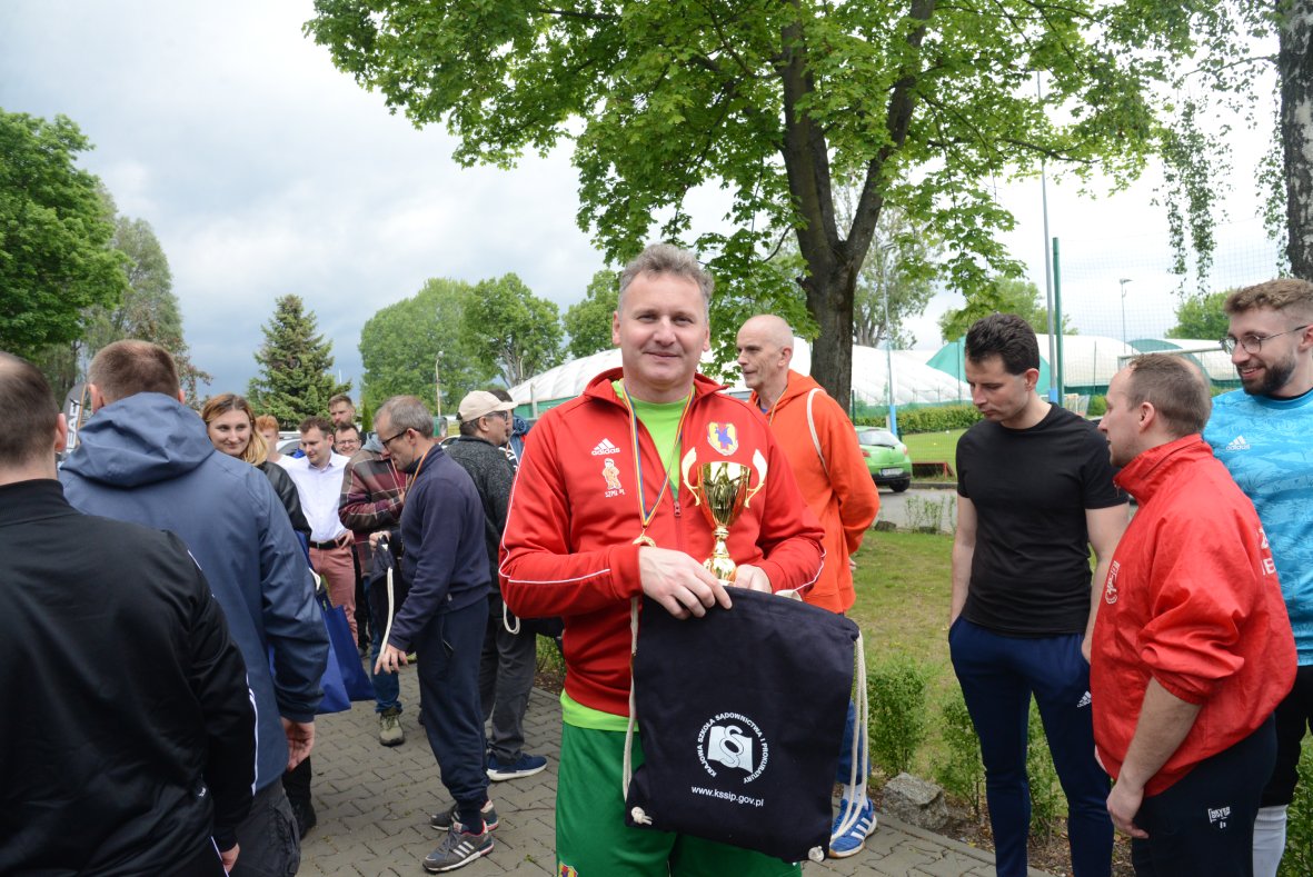 Na zdjęciu znajduje się grupa osób zgromadzonych na chodniku pod drzewami. Na pierwszym planie stoi mężczyzna w czerwonej bluzie sportowej z herbem i zielonych spodenkach, który trzyma w rękach złoty puchar oraz granatowy worek z białym logo i napisem „kssip.gov.pl”. Wokół niego stoją inni uczestnicy wydarzenia, niektórzy w sportowych strojach, inni w codziennych ubraniach. W tle widać drzewa, namioty oraz fragmenty boisk sportowych. 