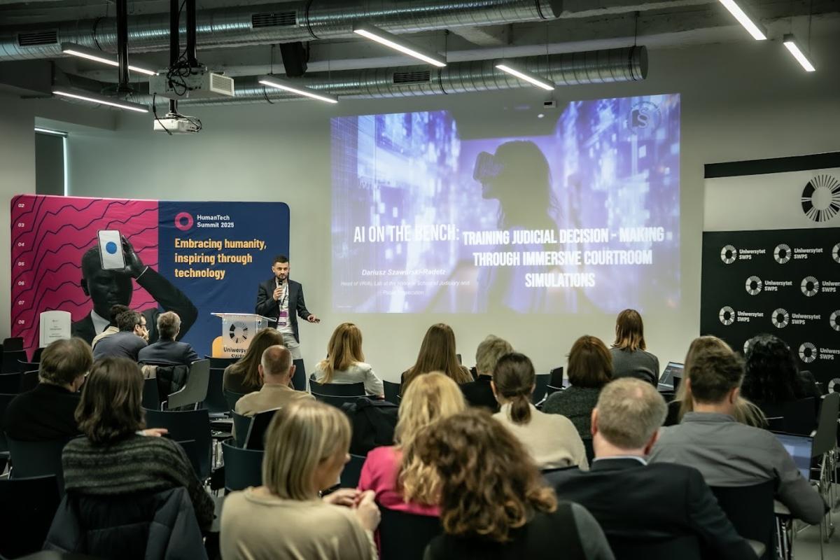 Zdjęcie przedstawia salę konferencyjną z grupą osób siedzących i słuchających prezentacji. Na pierwszym planie widać uczestników zwróconych w stronę ekranu. Na ekranie wyświetlona jest plansza z prezentacją zatytułowaną “AI on the Bench: Training Judicial Decision-Making Through Immersive Courtroom Simulations”. Prelegent stoi obok ekranu, trzyma mikrofon i mówi do publiczności. W tle widoczne są banery z logotypami wydarzenia i uczelni, a także hasło ‘Embracing humanity, inspiring through technology’. Pomieszczenie ma nowoczesny wystrój z widocznymi instalacjami sufitowymi.
