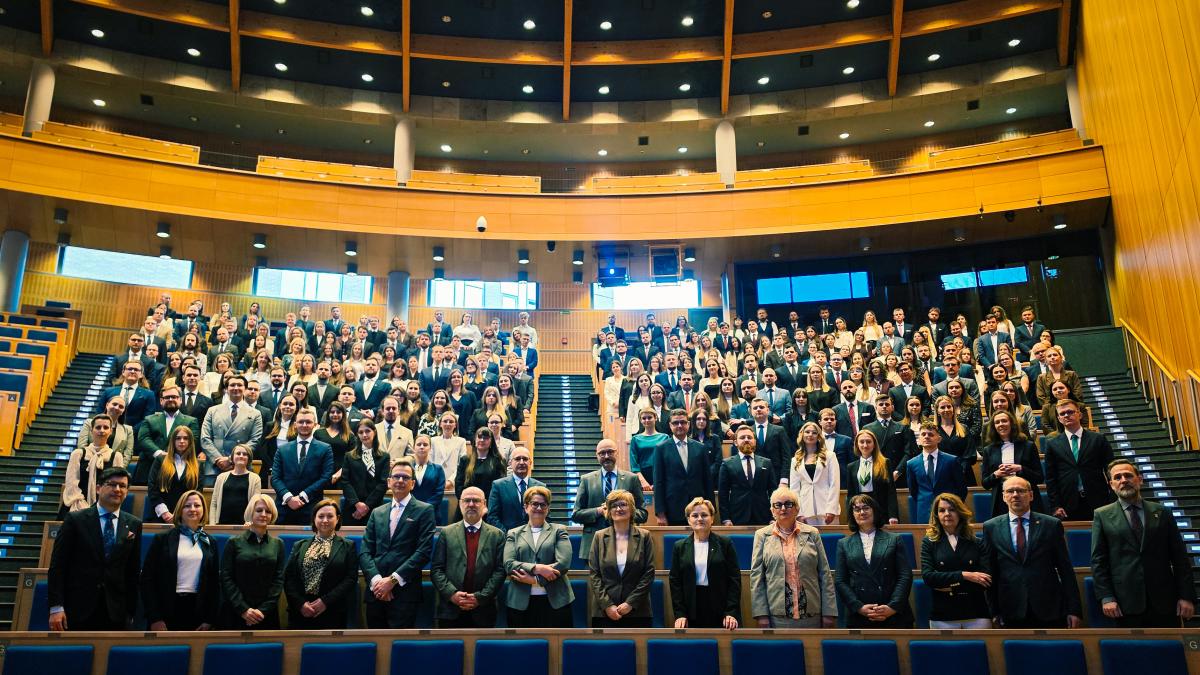 Na zdjęciu widać dużą grupę ludzi ustawionych w sali audytorium. Pomieszczenie jest przestronne, z półkolistym układem rzędów siedzeń, które wznoszą się ku górze. Ściany i elementy wnętrza są wykończone jasnym drewnem, a na suficie znajdują się liczne okrągłe światła. Osoby na zdjęciu stoją między rzędami siedzeń i są ubrane elegancko w garnitury, marynarki, koszule i sukienki. W najniższych dwóch rzędach, stoi kilka osób. Za nimi widać liczną grupę młodych ludzi rozłożonych symetrycznie po sali. Przez środek oraz po lewej i prawej stronie przebiegają schody. Wszyscy patrzą przed siebie.