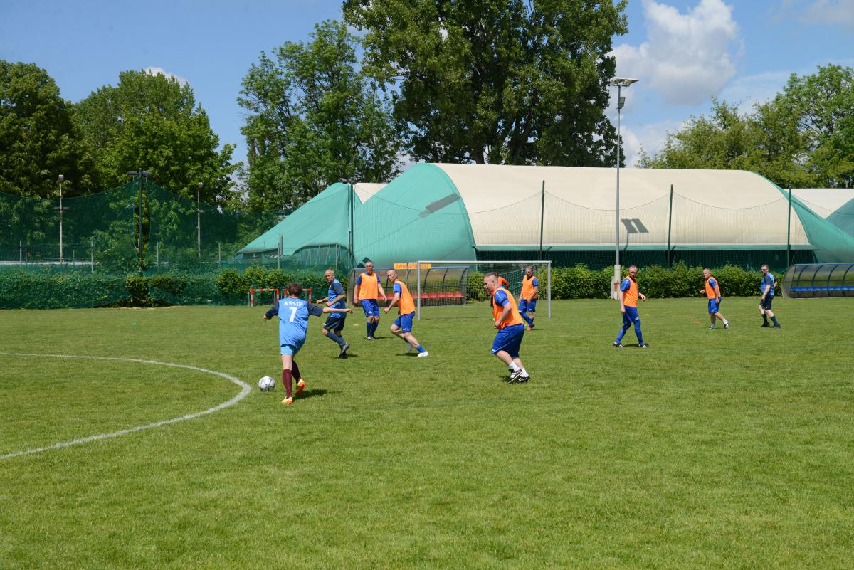 Zdjęcie nr: 24-DSC_3341 przedstawia odbywający się na murawie mecz piłkarski drużyn ubranych w granatowoczarne stroje oraz w stroje niebieskie. Zawodnicy w niebieskim stroju piłkarskim są dodatkowo ubrani w pomarańczową koszulkę na ramiączkach.