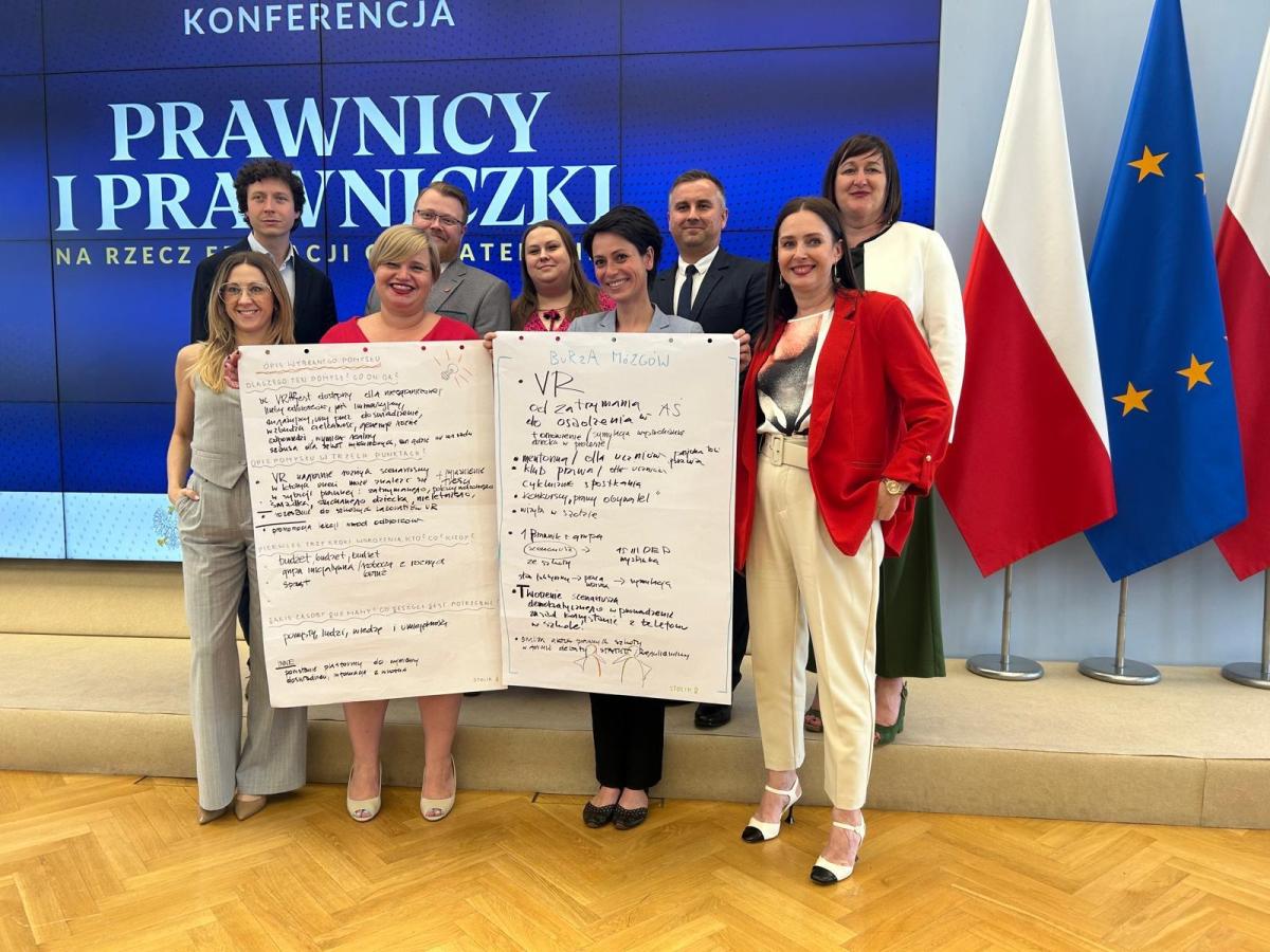 Grupa dziewięciu osób, zarówno kobiet, jak i mężczyzn, pozuje do zdjęcia na tle dużego ekranu z napisem „KONFERENCJA PRAWNICY I PRAWNICZKI NA RZECZ EDUKACJI OBYWATELSKIEJ”. Dwie osoby w środku trzymają duże arkusze papieru z odręcznymi notatkami. W tle widoczne są flagi Polski i Unii Europejskiej. Uczestnicy są uśmiechnięci i ubrani w eleganckie stroje.