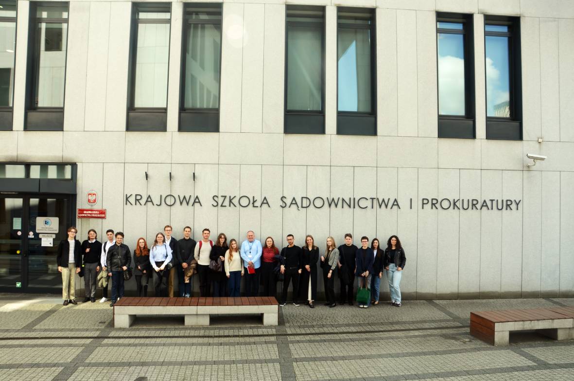 Grupa około dwudziestu osób stojących w szeregu przed nowoczesnym budynkiem z jasnoszarego kamienia. Nad nimi, na ścianie budynku, znajduje się duży napis: „Krajowa Szkoła Sądownictwa i Prokuratury”. Osoby są różnie ubrane – część elegancko, część swobodnie. Po lewej stronie widać wejście do budynku z przeszklonymi drzwiami, a przed grupą ustawiona jest betonowa ławka. W tle widać wysokie okna budynku.