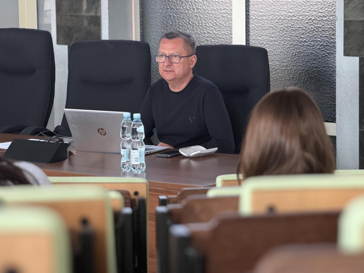 Mężczyzna w okularach siedzi przy stole konferencyjnym i prowadzi spotkanie. Ma na sobie ciemną koszulkę i pracuje na otwartym laptopie. Na stole znajdują się dwie butelki wody, telefon oraz komputerowa myszka. W tle widać wysokie, czarne krzesła oraz przeszkloną ścianę. Na pierwszym planie widoczni są uczestnicy siedzący w rzędach, zwróceni w stronę prowadzącego. Scena ma charakter formalny i szkoleniowy.