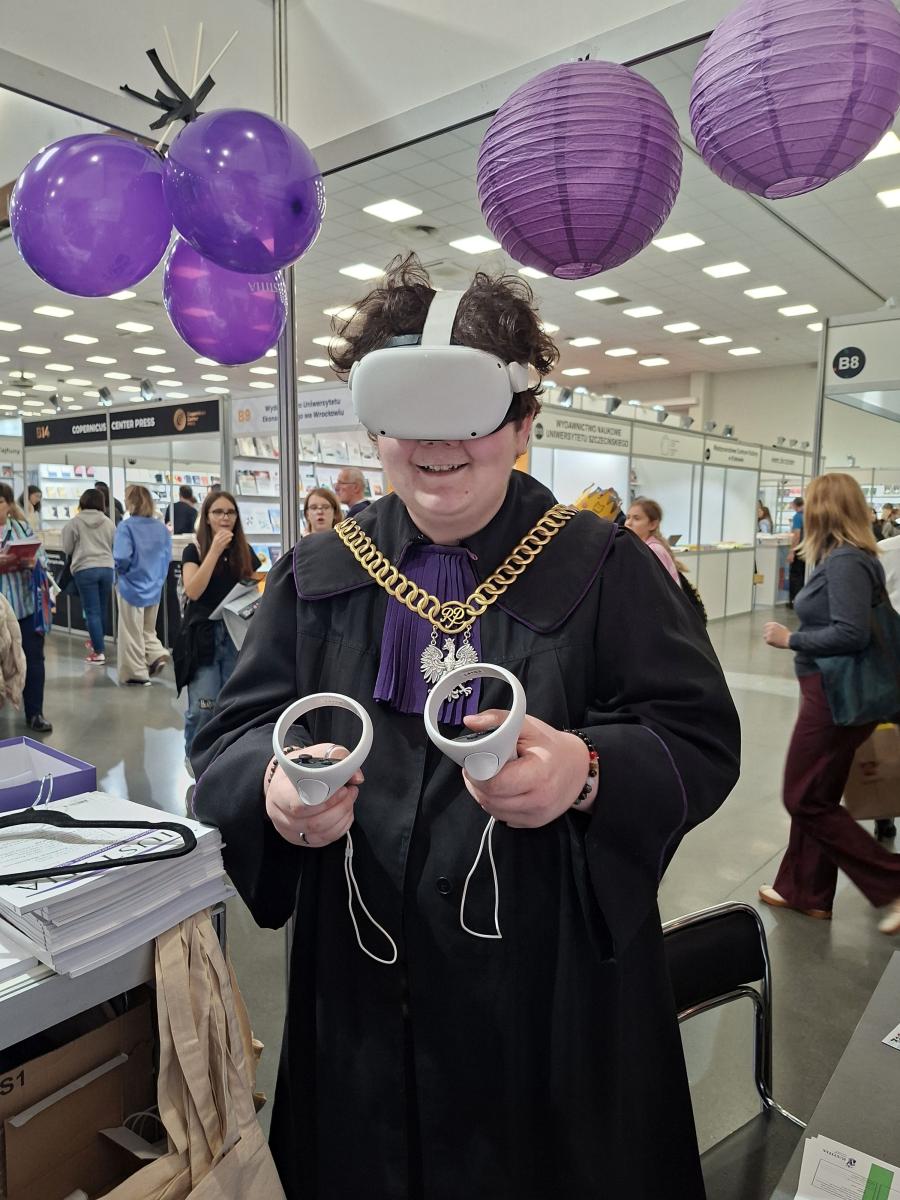 Na zdjęciu widać osobę w todze sędziowskiej z fioletowym żabotem i złotym łańcuchem z wizerunkiem orła, która testuje gogle wirtualnej rzeczywistości. Trzyma w dłoniach dwa kontrolery VR i uśmiecha się. Za nią widać stoisko targowe z odwiedzającymi, a przestrzeń udekorowana jest fioletowymi balonami i papierowymi lampionami. Zdjęcie oddaje atmosferę ciekawości i zabawy, pokazując połączenie tradycji sądownictwa z nowoczesną technologią.