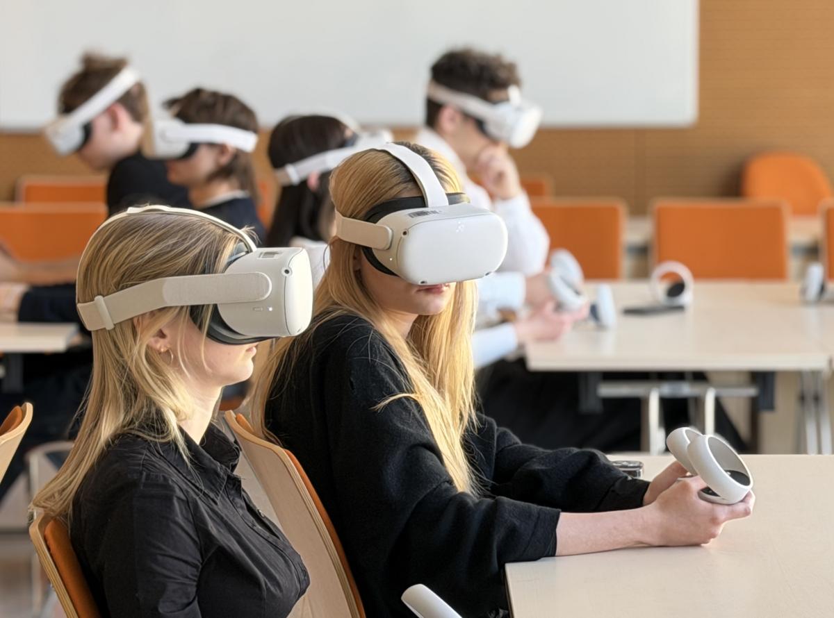Warsztaty VR w Pracowni VR/AI; Grupa młodych osób siedzi przy stołach w sali szkoleniowej. W pierwszym planie widoczne są dwie kobiety w ciemnych ubraniach, które mają na głowach gogle wirtualnej rzeczywistości i w dłoniach trzymają kontrolery. W tle siedzą kolejne osoby korzystające z gogli. Sala jest nowoczesna i jasna, z jasnymi ścianami oraz pomarańczowymi krzesłami.