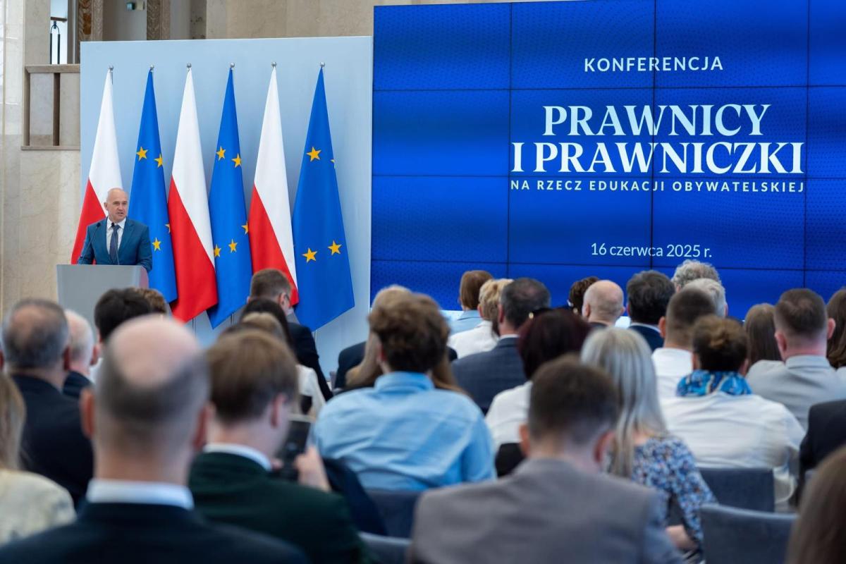 Widok z tyłu na salę konferencyjną, gdzie uczestnicy siedzą zwróceni w stronę sceny. Na scenie przy mównicy stoi mężczyzna w garniturze, przemawiający do publiczności. W tle duży ekran z napisem „KONFERENCJA PRAWNICY I PRAWNICZKI NA RZECZ EDUKACJI OBYWATELSKIEJ”, a poniżej data „16 czerwca 2025 r.”. Po obu stronach sceny stoją flagi Polski i Unii Europejskiej.