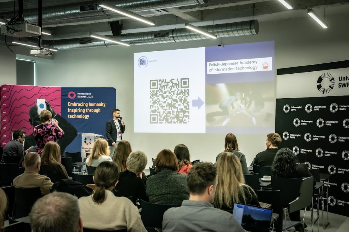 Zdjęcie przedstawia salę konferencyjną z grupą osób siedzących i słuchających prezentacji. Na pierwszym planie widać uczestników zwróconych w stronę ekranu. Na ekranie wyświetlony jest duży kod QR po lewej stronie oraz napis „Polish-Japanese Academy of Information Technology” po prawej stronie, obok grafiki z osobami w ruchu. Prelegent stoi obok ekranu, trzyma mikrofon i mówi do publiczności. W tle widoczne są banery z logotypami wydarzenia i uczelni, a także hasło „Embracing humanity, inspiring through technology”. Pomieszczenie ma nowoczesny wystrój z widocznymi instalacjami sufitowymi.