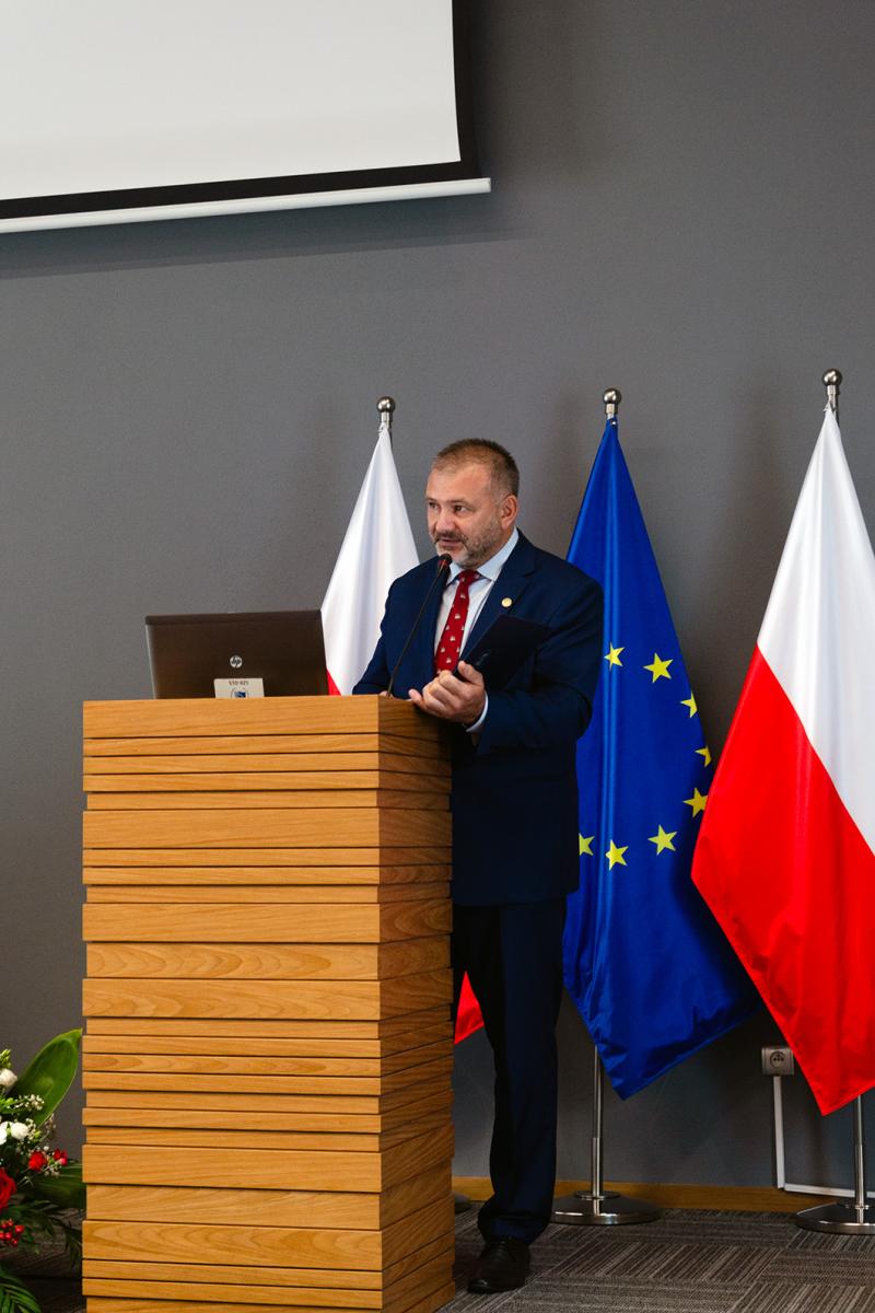 Przedstawia mężczyznę – Dyrektora ds. Organizacyjnych Krajowej Szkoły Sądownictwa  i Prokuratury, Sędziego Waldemara Żurka. Mężczyzna ubrany w granatowy garnitur, białą koszulę i czerwony krawat znajduje się za drewnianą mównicą w naturalnym kolorze jasnego drewna. Przed nim na mównicy stoi laptop, za nim znajduje się flaga Unii Europejskiej oraz dwóch flag Polski, znajdujących się po obu jej stronach. Wszystkie flagi umieszczone są na stojaku flagowym. Nad mężczyzną widać fragment ekranu do rzutnika. Obok mównicy widać fragment wiązanki kwiatowej w kolorze biało-czerwonym. Zdjęcie wykonane jest na tle szarej ściany. Mężczyzna trzyma w ręce okładkę, w której znajdują się notatki do wygłaszanego przemówienia. 