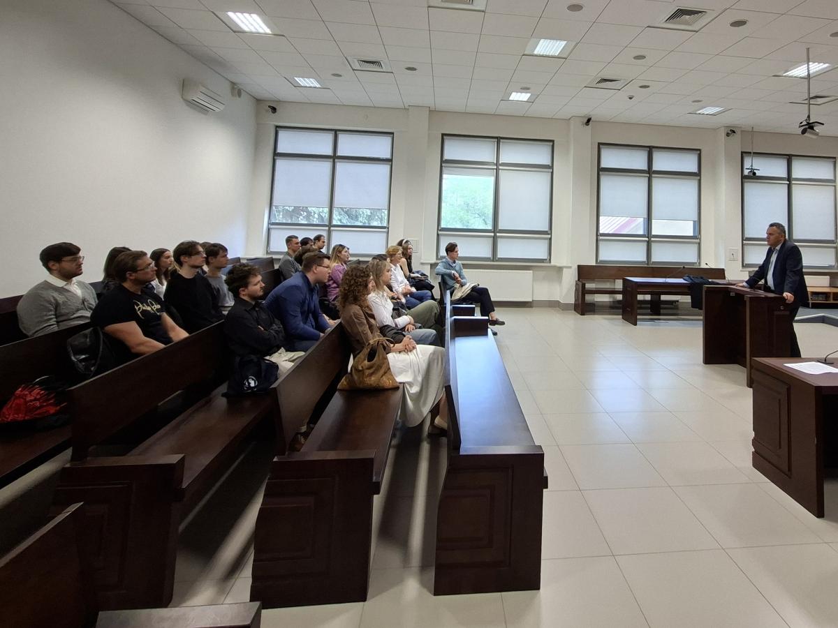 Wnętrze jasnej sali sądowej. Na pierwszym planie po lewej stronie siedzi grupa młodych osób. Po prawej stronie stoi mężczyzna w garniturze. W tle widać ławy, okna z opuszczonymi roletami oraz sprzęt nagrywający na suficie.