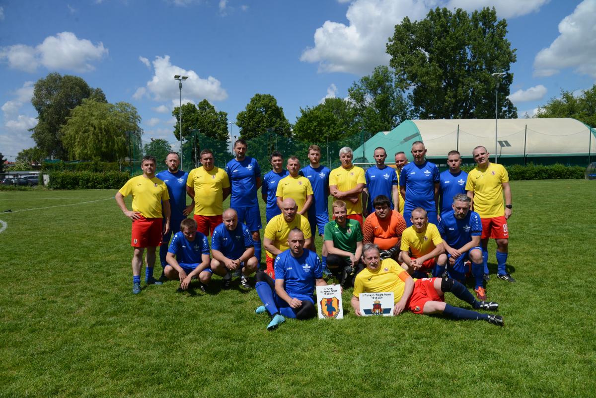 Zdjęcie nr: 8-DSC_3329 przedstawia wspólne skład dwóch drużyn piłkarskich - łącznie dziewiętnaście mężczyzn, na murawie piłkarskiej, ośmiu z nich ubranych jest strój piłkarski koloru  żółtoczerwonego,  jedenastu w strój koloru niebieskiego, jeden w strój bramkarski koloru zielonego, jeden w strój bramkarski koloru pomarańczowego. Trzynastu  mężczyzn stoi, siedmiu kuca, dwóch spoczywa na trawie trzymając w rękach logo drugiej drużyny .