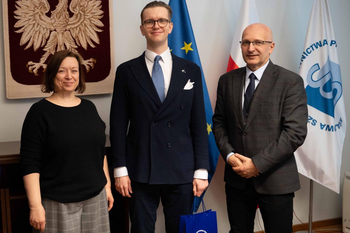 Na zdjęciu widać trzy osoby stojące obok siebie w pomieszczeniu. Po lewej stronie stoi kobieta z krótkimi, ciemnymi włosami. Ubrana jest w czarną bluzkę oraz szerokie, szare spodnie w kratę. Ma lekki uśmiech. Po środku stoi młody mężczyzna w eleganckim, granatowym garniturze z krawatem i białą poszetką. Ma okulary, zaczesane włosy w tył i lekko się uśmiecha. W ręce trzyma niebieską torbę. Po prawej stronie stoi mężczyzna w ciemnym garniturze, koszuli i krawacie, z rękami złożonymi przed sobą. Ma okulary i lekko się uśmiecha. Na ścianie za nimi widoczny jest biały orzeł na czerwonym tle – symbol państwowy Polski, oraz trzy flagi na stojakach: flaga Unii Europejskiej (niebieska z żółtymi gwiazdami), flaga Polski (biało-czerwona) i flaga Krajowej Szkoły (biała z niebieskimi napisami i symbolem paragrafu). Ściana za osobami jest jasna biała.