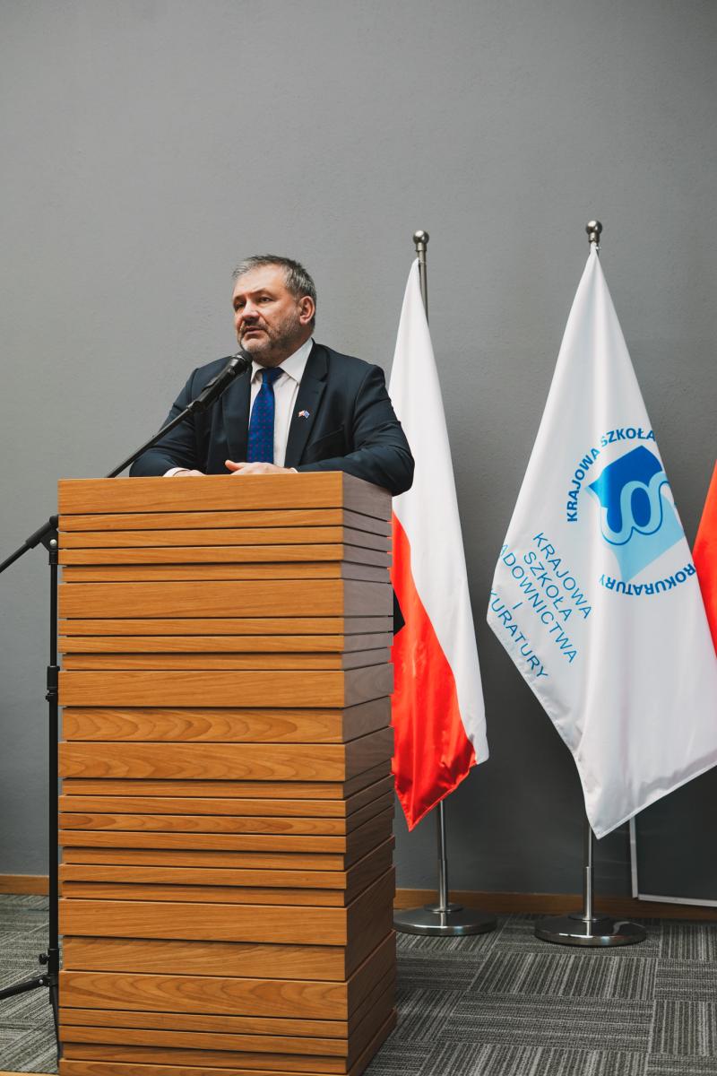 Na zdjęciu widoczny jest mężczyzna stojący przy mównicy, który wygłasza oficjalne przemówienie. Ubrany jest w elegancki, formalny strój: ciemną marynarkę, jasną koszulę oraz niebieski krawat, co podkreśla rangę wydarzenia. Jego postawa jest skupiona, rękami opiera się o mównicę, a uwaga skierowana jest przed siebie. W tle widoczna jest jednolita, szara ściana nadająca scenie stonowany, profesjonalny charakter. Po prawej stronie ustawione są flagi — flaga Polski oraz flaga z logo Krajowej Szkoły Sądownictwa i Prokuratury, co sugeruje, że wydarzenie ma charakter oficjalny.