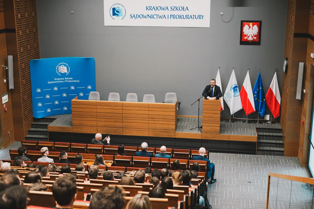 Na zdjęciu przedstawiona jest duża, przestronna sala przypominająca aulę lub salę wykładową. W pomieszczeniu zgromadzonych jest wiele osób siedzących w równych, uporządkowanych rzędach. Uczestnicy wydarzenia zwróceni są plecami do fotografa i skierowani w stronę przodu sali, gdzie znajduje się scena z mównicą. Przy mównicy stoi mężczyzna wygłaszający przemówienie. Ubrany jest w oficjalny strój składający się z marynarki, koszuli oraz krawata, co podkreśla formalny charakter wydarzenia. Za jego plecami widoczne są flagi Polski, flaga Unii Europejskiej, flaga z logo Krajowej Szkoły Sądownictwa i Prokuratury oraz baner z logo Krajowej Szkoły Sądownictwa i Prokuratury. Obok mównicy ustawione są krzesła, jednak pozostają one puste. W sali panuje porządek i cisza, a atmosfera wydaje się skupiona i poważna. Wysoki sufit potęguje wrażenie przestronności, a na ścianie za mównicą widoczne jest godło z wizerunkiem orła, co dodatkowo wskazuje na oficjalny, instytucjonalny charakter wydarzenia.