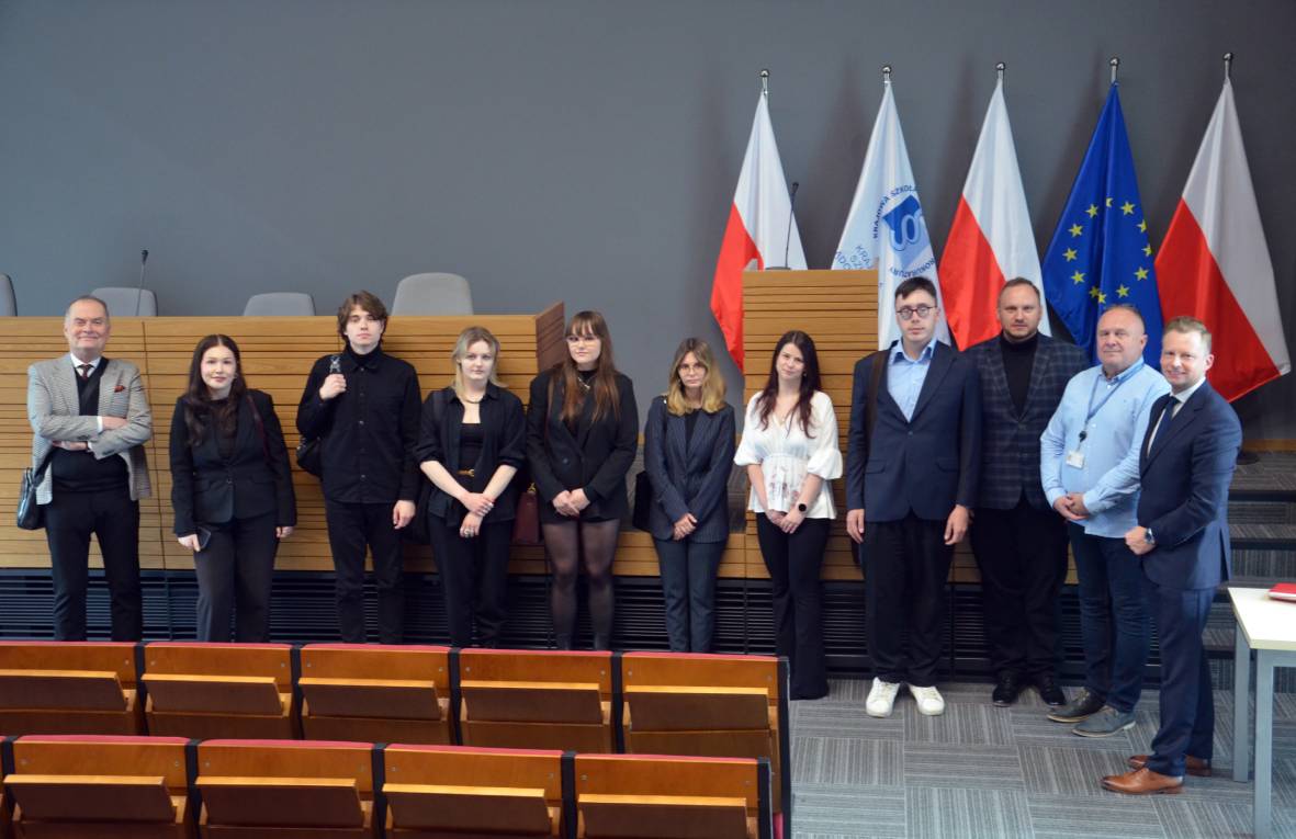 Na zdjęciu grupa jedenastu osób stoi obok siebie. Osoby znajdują się w sali z drewnianymi ławkami i podwyższeniem z miejscami dla prezydium. W tle znajdują się trzy polskie flagi, jedna flaga Unii Europejskiej i jedna flaga Krajowej Szkoły Sądownictwa i Prokuratury. 