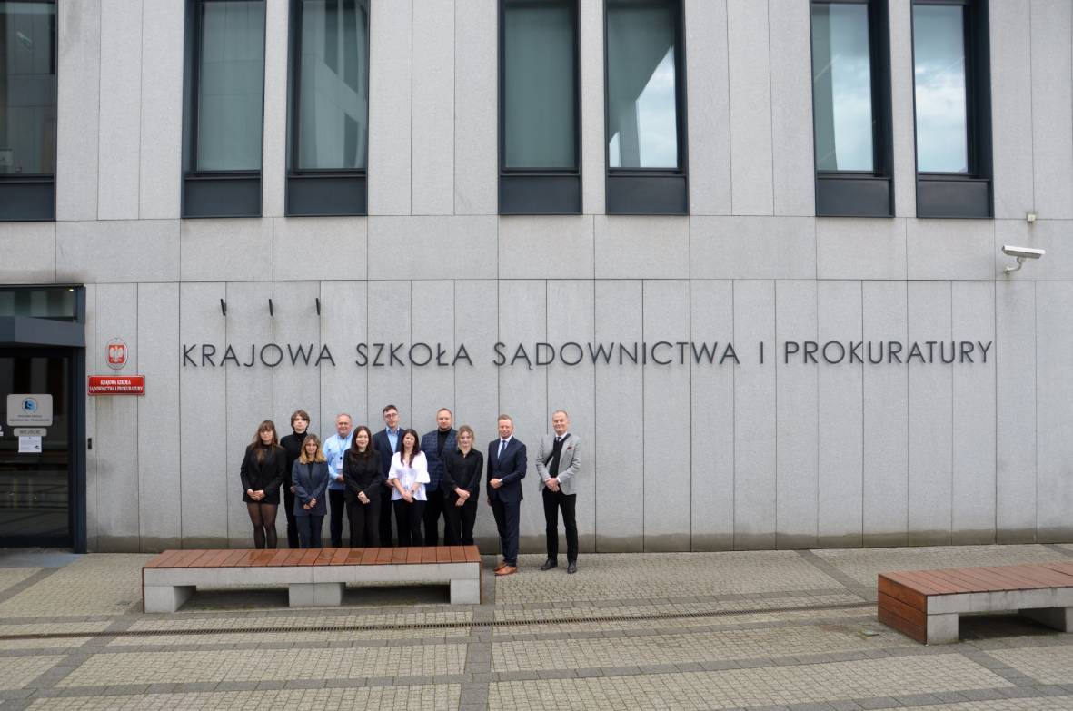 Grupa jedenastu osób stojących przed nowoczesnym, jasnoszarym budynkiem. Na ścianie budynku znajduje się duży napis: „Krajowa Szkoła Sądownictwa i Prokuratury”. Po lewej stronie znajduje się wejście do budynku oraz czerwona tablica z nazwą Krajowej Szkoły. Przed grupą ustawione są dwie ławki. 