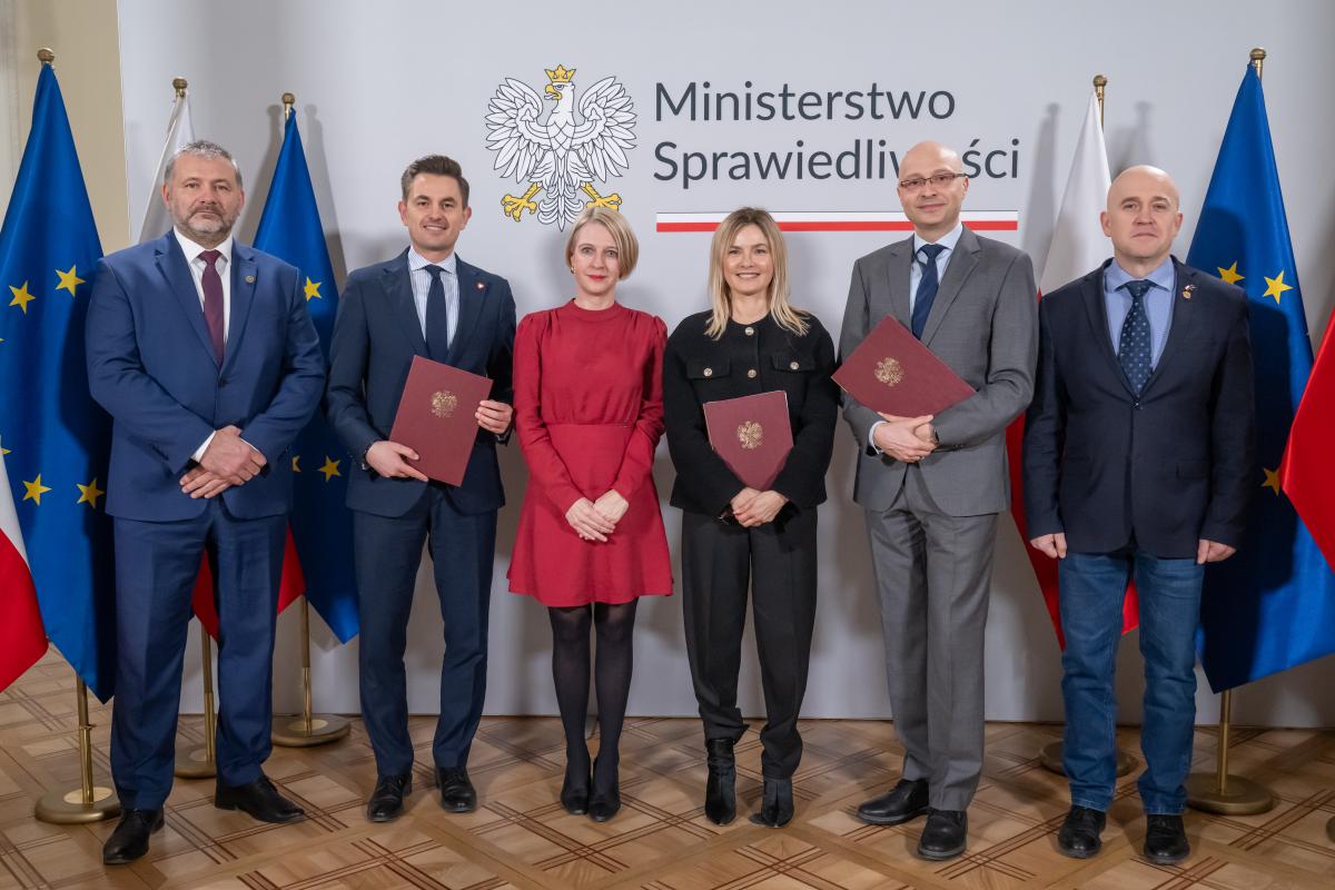 Czterech mężczyzn oraz dwie kobiety stoją na tle ściany z napisem Ministerstwo Sprawiedliwości. Dwóch mężczyzn oraz jedna kobieta trzymają w ręku brązowe teczki ze złotym orłem. Po obu stronach znajdują się flagi Polski i Unii Europejskiej.