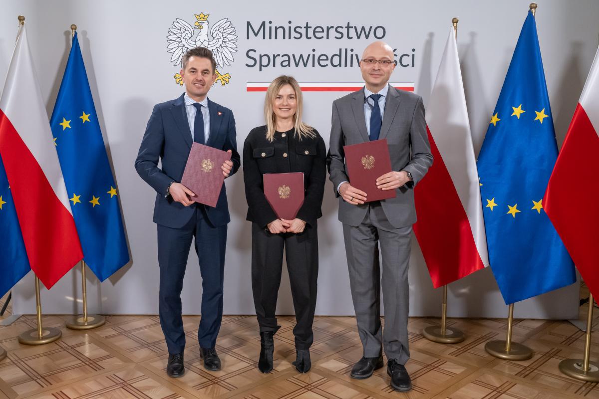 Dwóch mężczyzn oraz jedna kobiety stoją na tle ściany z napisem Ministerstwo Sprawiedliwości. W ręku trzymają brązowe teczki ze złotym orłem. Po obu stronach znajdują się flagi Polski i Unii Europejskiej.