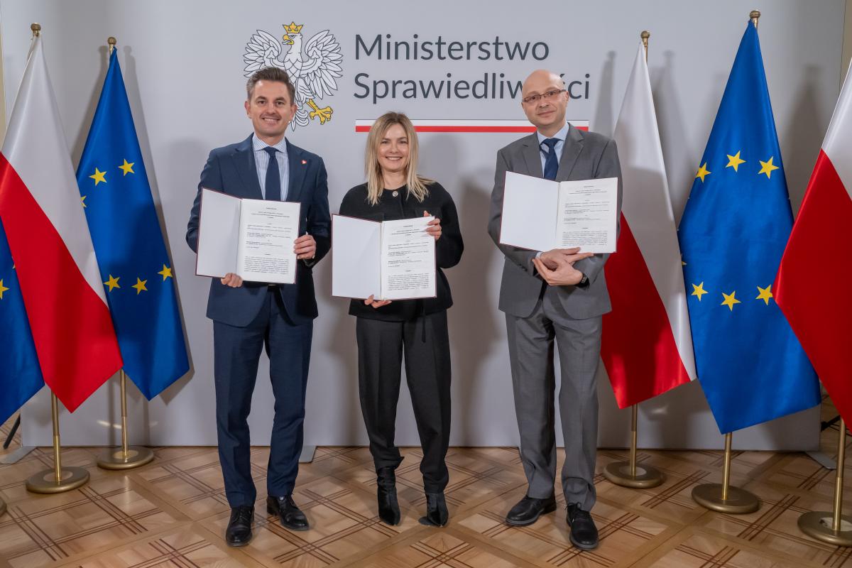 Dwóch mężczyzn oraz jedna kobiety stoją na tle ściany z napisem Ministerstwo Sprawiedliwości. W ręku trzymają otwarte teczki. Po obu stronach znajdują się flagi Polski i Unii Europejskiej.