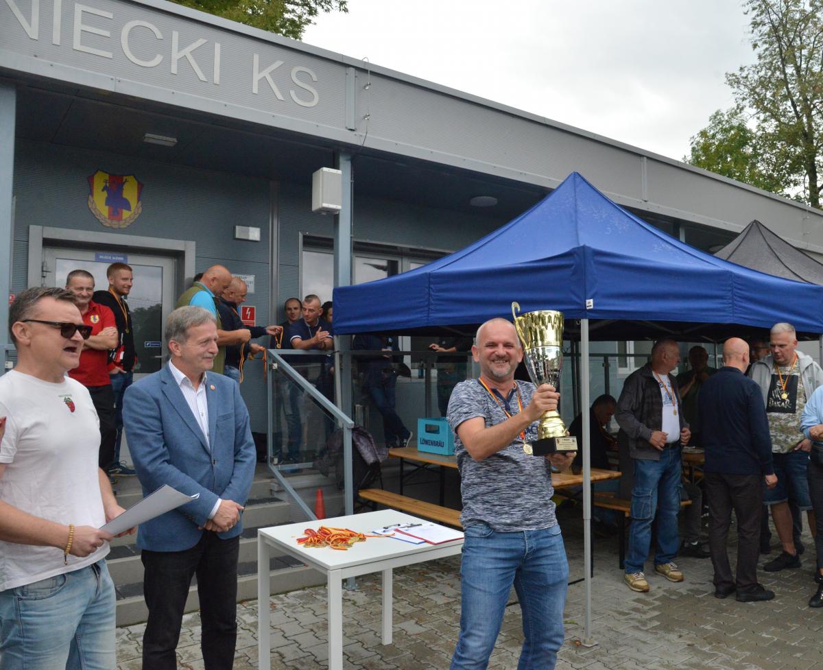 trzech mężczyzn na tle grupy osób, jeden z mężczyzn ma na sobie złoty medal i trzyma uniesiony puchar