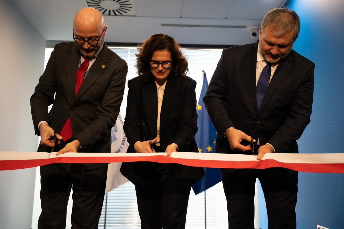 Wewnątrz pomieszczenia widoczne trzy osoby, dwóch mężczyzn i kobieta w środku. Osoby ubrane są w ciemne garnitury i białe koszule. Każda z widocznych osób równocześnie przecina biało-czerwoną wstęgę trzymanymi w ręku nożyczkami.