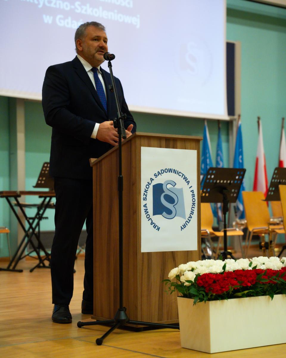 Wewnątrz pomieszczenia widoczny mężczyzna w ciemny garniturze, białej koszuli i granatowym krawacie. Mężczyzna stoi za mównicą z mikrofonem, przed którą stoją na podłodze biało-czerwone kwiaty. W tle widoczny u góry ekran, a po prawej stronie flagi oraz krzesła.