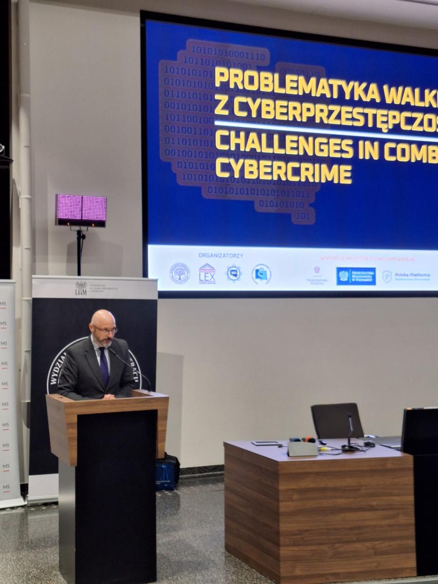 zdjęcie przedstawia mężczyznę przy mównicy w tle widoczny ekran na którym wyświetlony jest tytuł konferencji