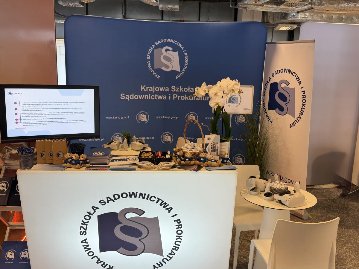 Na zdjęciu widać ladę wystawową, na której rozłożono różne gadżety z logo Krajowej Szkoły Sądownictwa i Prokuratury: pluszowe misie, notesy, długopisy, broszury, magnesy oraz słodkie krówki. Obok stoją stoliki wyposażone w gogle VR, a na ladzie znajduje się również monitor wyświetlający prezentację. W tle widoczny jest baner z logo oraz dekoracyjna roślina.