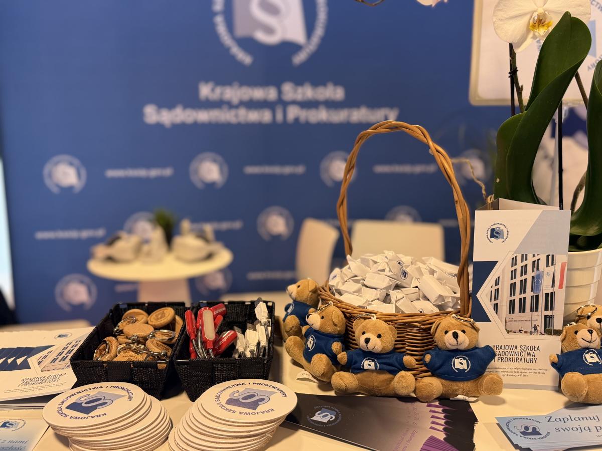 Na zdjęciu widać różne gadżety z logo — podkładki, breloki, zakładki oraz pluszowe misie. Na pierwszym planie znajduje się także koszyk ze słodkimi krówkami przeznaczonymi do poczęstunku. Tło jest rozmyte, ale można dostrzec baner z logo. Całe ujęcie jest wyraźnie sfokusowane na pierwszy plan, co podkreśla eksponowane przedmioty.
