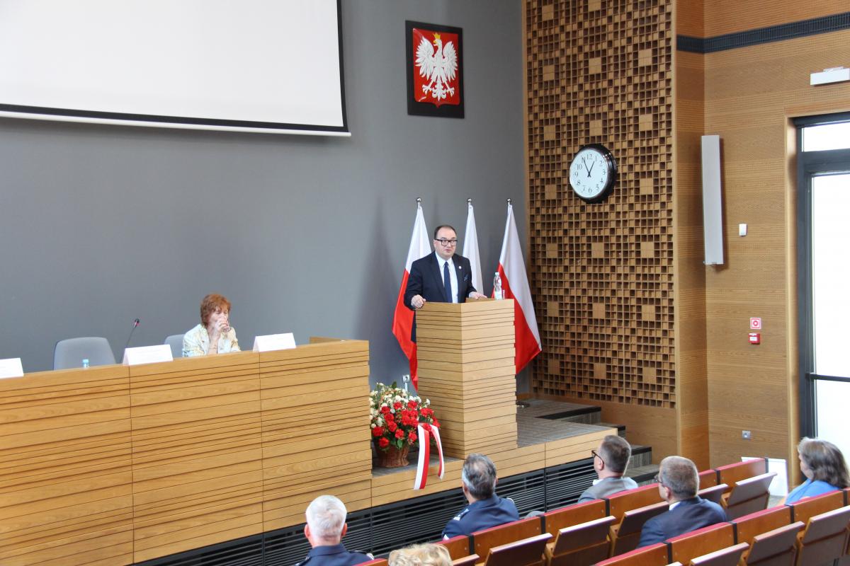 W auli KSSiP kobieta  siedzi za stołem prezydialnym, mężczyzna przemawia,  pozostałe osoby stoją.