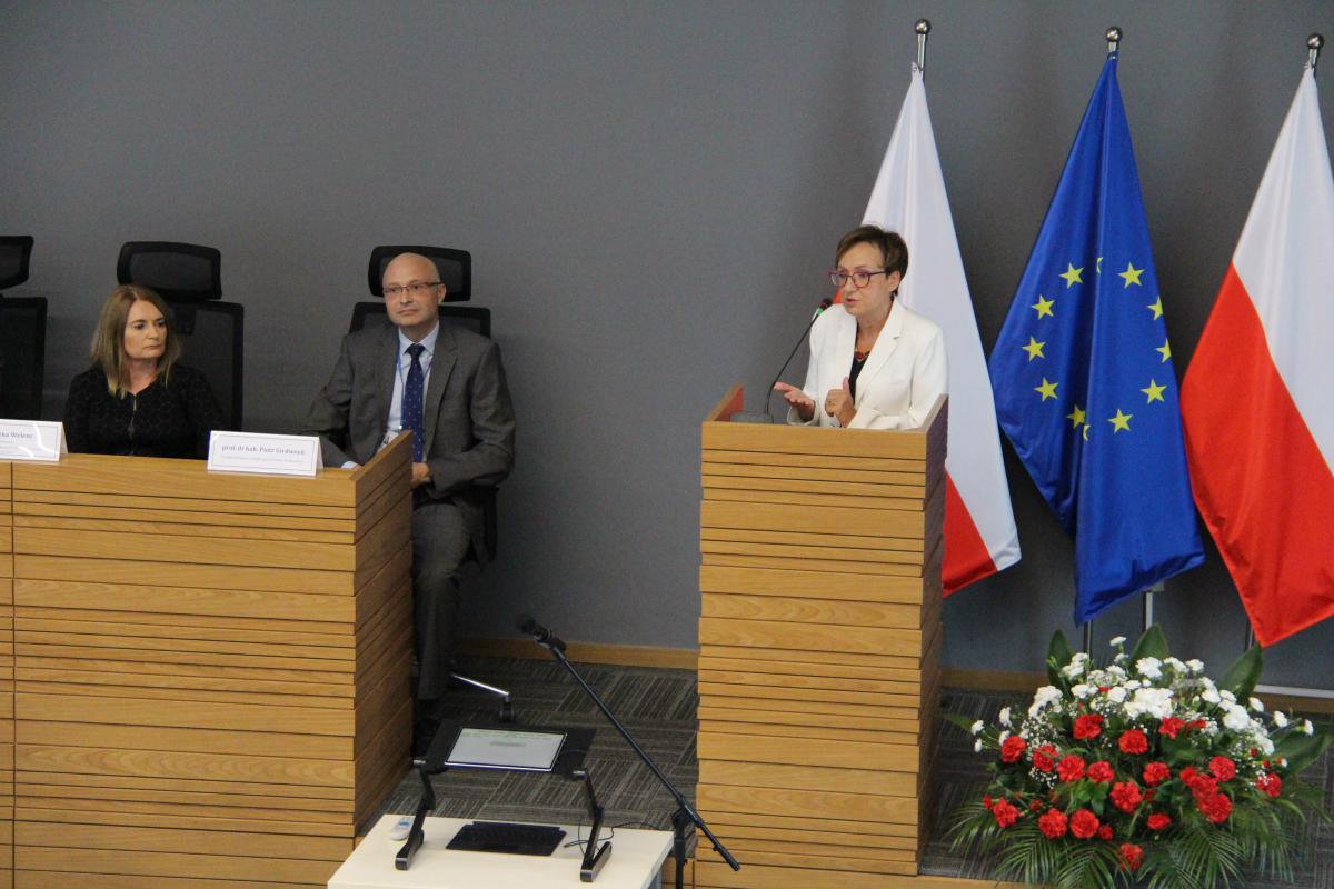 przedstawia przemawiająca kobietę, na tle dwóch flag w barwach narodowych: białej i czerwonej oraz flagi UE, obok za stołem z lewej strony siedzi kobieta i mężczyzna w okularach.