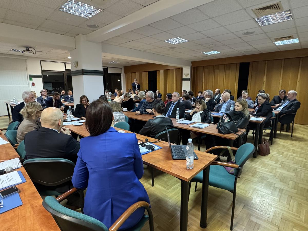 zdjęcie przedstawia salę konferencyjną z rozstawionymi czteroosobowymi stołami, za którymi siedzi kilkadziesiąt osób, obecni zwróceni są twarzami w stronę stojącego przed oknem, przemawiającego do mikrofonu mężczyzny.