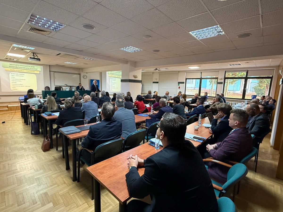 zdjęcie przedstawia salę konferencyjną z rozstawionymi czteroosobowymi stołami, za którymi siedzi kilkadziesiąt kobiet i mężczyzn, twarzami zwróconymi w kierunku stołu konferencyjnego. Za stołem konferencyjnym siedzi mężczyzna, przed nim stoi przemawiający mężczyzna. Na ścianie znajdującej się za stołem konferencyjnym, naprzeciwko słuchaczy, wyświetlana jest prezentacja pn.„Metody dydaktyczne wykorzystywane w KSSiP”;