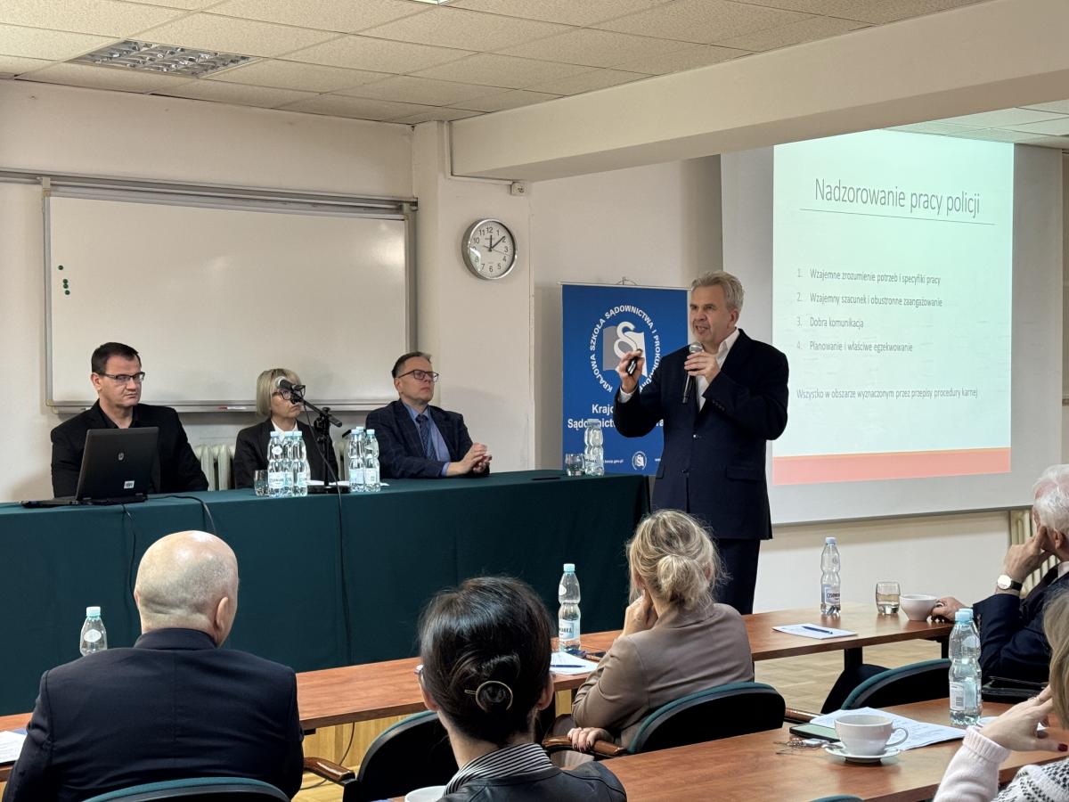 zdjęcie przedstawia przemawiającego mężczyznę, trzymającego w rękach mikrofon i pilot do projektora. Za przemawiającym mężczyzną znajduje się stół konferencyjny, przy którym siedzi kobieta oraz dwóch mężczyzn. Za nimi na ścianie wyświetlany jest slajd pn. „nadzorowanie pracy policji”. Przed stołem konferencyjnym widać stoły, za którym siedzi kilka osób, mężczyźni i kobiety, z twarzami zwróconymi w kierunku mówcy. 