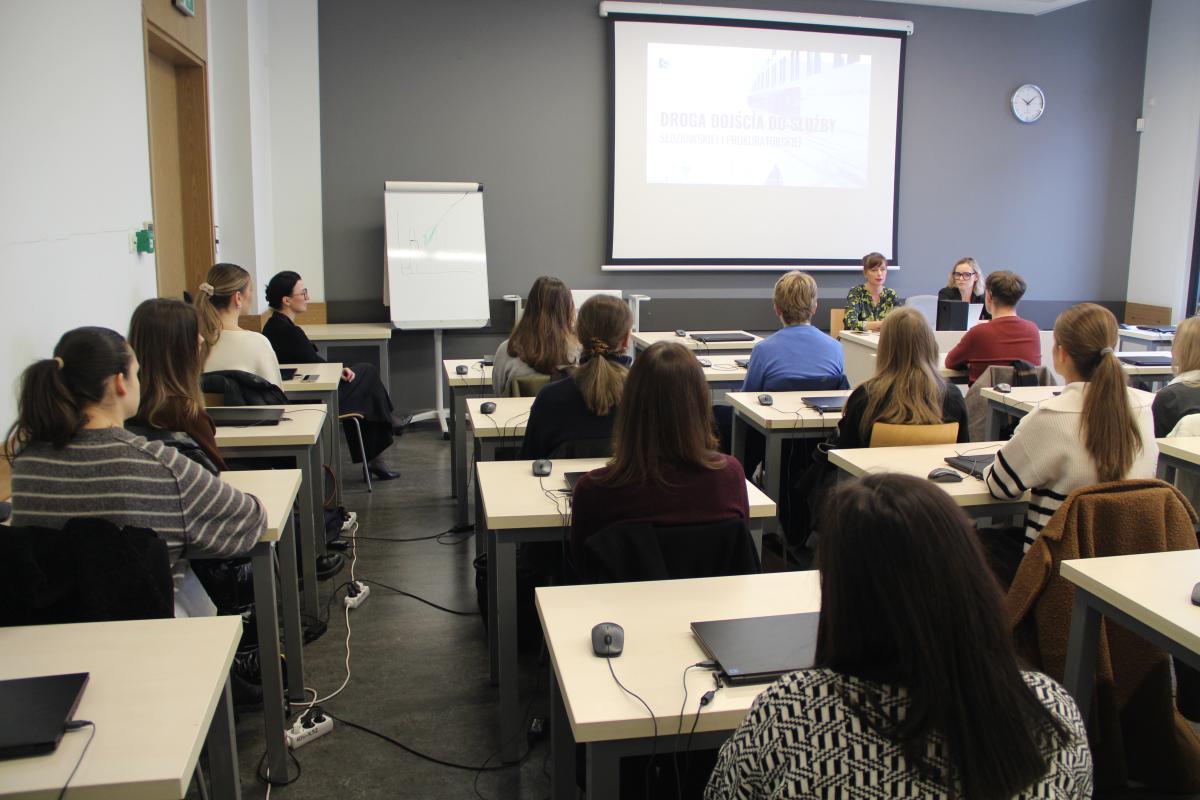 Grupa mężczyzn i kobiet siedzi przy stolikach, tyłem do obiektywu aparatu. Na większości stolików widoczne są laptopy. Naprzeciwko grupy kobiet i mężczyzn ustawione jest biurko z laptopem, przy którym siedzą 2 kobiety. W tle widać rzutnik z prezentacją: Droga dojścia do służby sędziowskiej i prokuratorskiej. Po lewej stronie, w rogu stoi duża biała tablica.