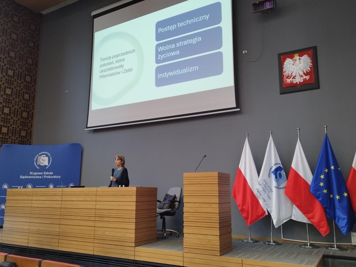 Zdjęcie przedstawia część auli wykładowej. Na pierwszym planie, za mównicą, widoczny jest mężczyzna w ciemnym garniturze, białej koszuli i bordowym krawacie. Po prawej stronie zdjęcia znajdują się, ustawione w rzędzie, flagi: Polski, Unii Europejskiej oraz Krajowej Szkoły. W oddali, po lewej stronie, za katedrą, znajduje się baner w kolorze granatowym z napisem Krajowa Szkoła Sądownictwa i Prokuratury oraz logiem Krajowej Szkoły.