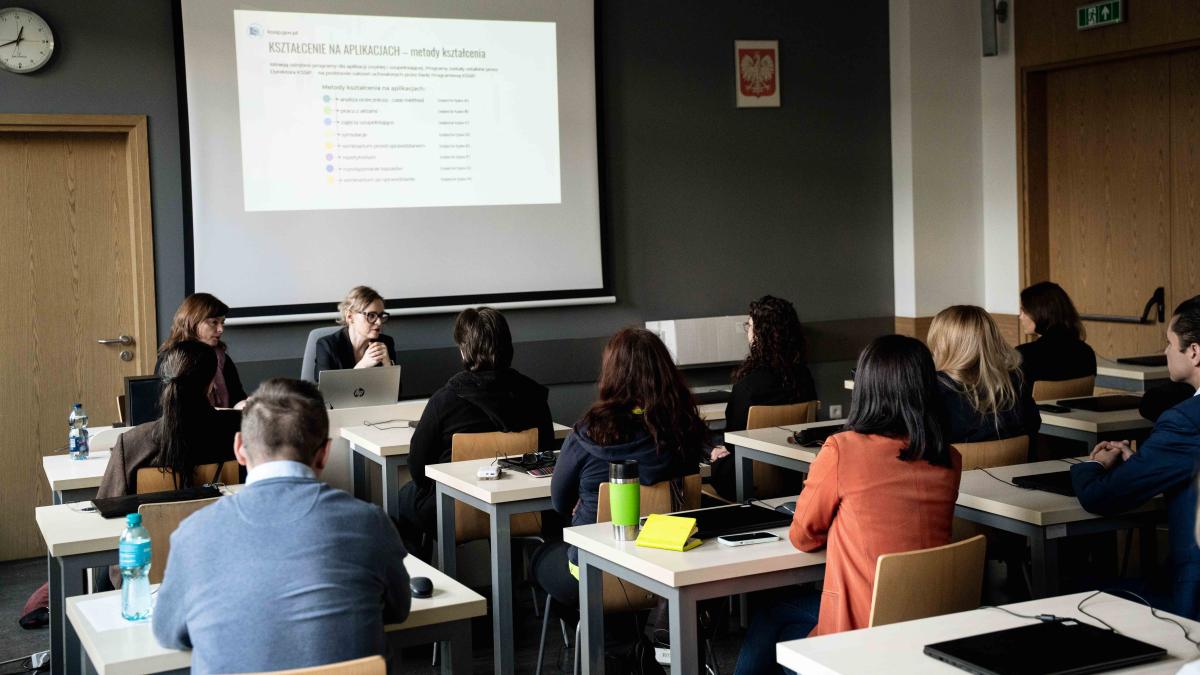 grupa 11 osób (kobiet i mężczyzn) siedzi przy stolikach. Dwie kobiety siedzą zwrócone twarzą do obiektywu. Przed nimi na stole stoi laptop i butelka wody. Na nimi wisi prezentacja dotycząca metod kształcenia na aplikacjach. Pozostałe osoby siedzą tyłem do obiektywu. Na ścianie po prawej stronie wisi godło Polski