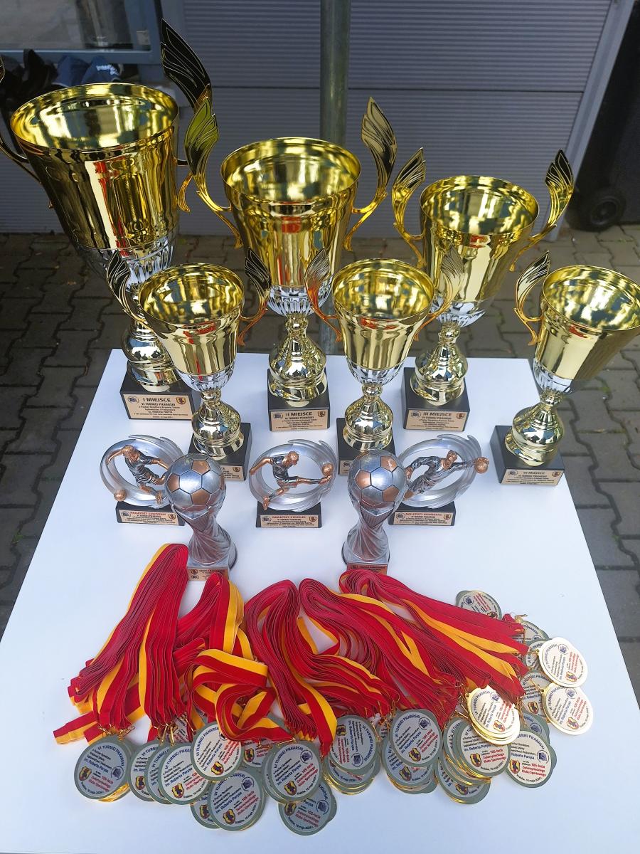 Zdjęcie zostało zrobione z góry. Na zdjęciu znajduje się biały stół z nagrodami sportowymi. Pięć złotych pucharów ustawionych w dwóch rzędach. Trzy puchary różnią się wielkością od pozostałych, ponieważ są przeznaczone dla drużyn, które zdobędą pierwsze, drugie i trzecie miejsce. Przed pięcioma złotymi pucharami znajdują się trzy srebrne statuetki przedstawiające sylwetę piłkarza wpisaną w okrągły element dekoracyjny. Są to nagrody dla najlepszego zawodnika, najlepszego strzelca i najlepszego bramkarza Turnieju. Przed nagrodami indywidualnymi znajdują się dwie srebrne statuetki, których elementem dekoracyjnym jest piłka do gry w piłkę nożną. Te statuetki są przeznaczone dla sędziów sędziujących w Turnieju. Na samym przodzie stołu leżą medale przeznaczone dla wszystkich zawodników biorących udział w Turnieju. Medale są okrągłe, złote, mają wyklejki z grafiką i tekstami oraz wstążki w czerwono-żółte pasy. W tle widoczna jest nawierzchnia z kostki brukowej, na której stoi stół.