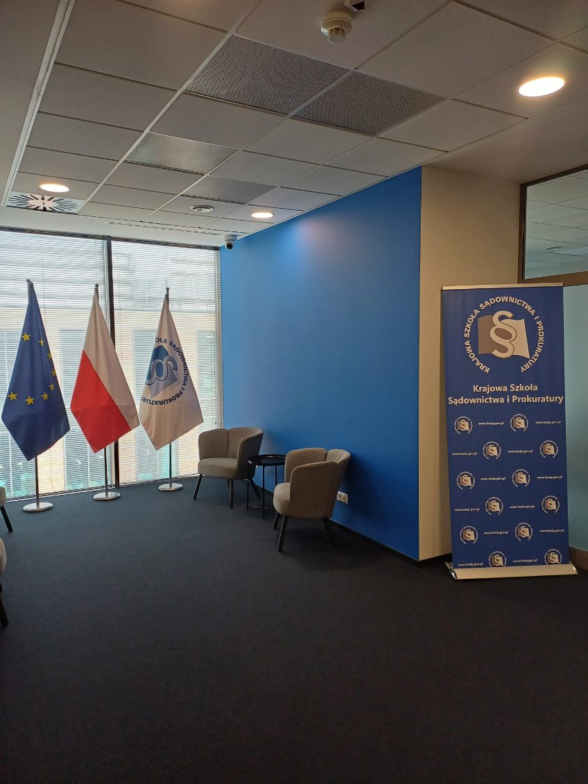 Przedstawia pusty hol w  nowej siedzibie KSSiP w Gdańsku. Na pierwszym planie wykładzina, w głębi od lewej – na tle okna – trzy flagi: Unii Europejskiej, Rzeczypospolitej Polskiej, Krajowej Szkoły, dwa fotele i stolik kawowy, flipchart Krajowej Szkoły. 