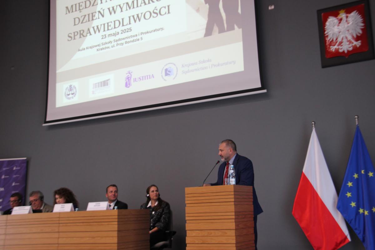 Trzech mężczyzn i dwie kobiety siedzą przy drewnianym pulpicie, na którym ustawione są białe tabliczki. Obok, przy mównicy, stoi mężczyzna. W tle znajduje się godło Polski oraz duży slajder z wyświetloną prezentacją z napisem Międzynarodowy Dzień Wymiaru Sprawiedliwości 23 maja 2025 r. 