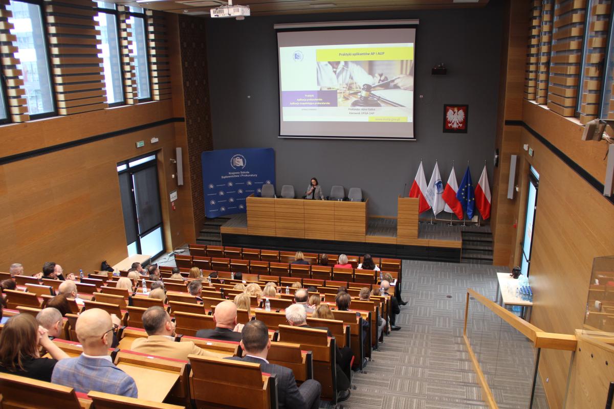 Fotografia przedstawia widok auli wykładowej. Na pierwszym planie widoczne są (od tyłu) liczne osoby zasiadające w fotelach. W dalszej perspektywie, na środku fotografii, znajduje się postać kobiety stojącej za katedrą. Kobieta ma ciemne włosy. Ubrana jest w marynarkę szarym kolorze i czarną bluzkę. Za kobietą, trzymającą w dłoni mikrofon, znajduje się ekran, na którym wyświetlana jest prezentacja.