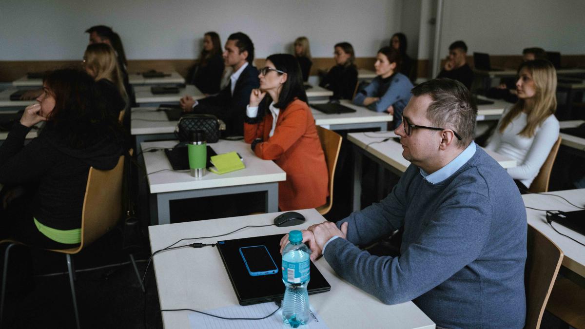 grupa 14 osób (kobiet i mężczyzn) siedzi przy stolikach. Na stolikach leżą laptopy. Na pierwszym planie widoczny jest mężczyzna w okularach. Na jego stoliku, na laptopie leży telefon komórkowy. Obok stoi mała butelka wody