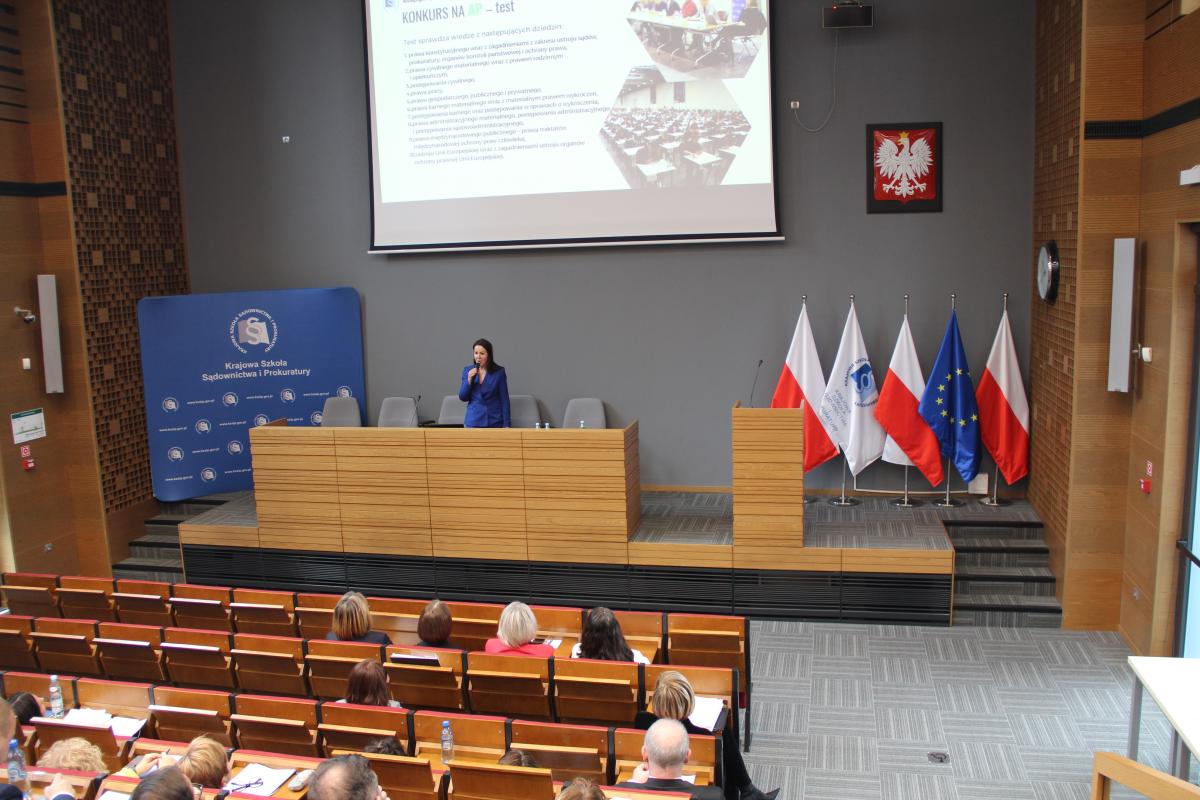 Zdjęcie przedstawia widok od środka auli wykładowej w kierunku katedry. Na zdjęciu, w jego centralnej części, widoczna jest kobieta o ciemnych długich włosach, ubrana w kobaltowy garnitur. Kobieta stoi za katedrą, trzyma w dłoni mikrofon. Za kobietą widoczny jest, na tle szarej ściany, ekran z wyświetlaną prezentacją.