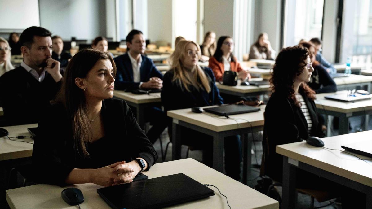 grupa ok. 16 osób (kobiet i mężczyzn) siedzi przy stolikach. Na stolikach leżą laptopy. Po prawej stronie znajdują się duże jasne okna.Twarze osób na zdjęciu są częściowo niewyraźne